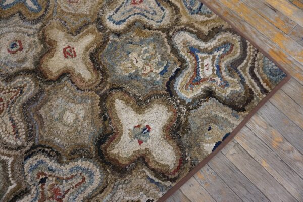 Close-up of a textured, multi-toned hooked rug featuring abstract star or cross patterns in blues, browns, and creams, resting on rustic wooden floorboards by rugs on net. By rugs on net