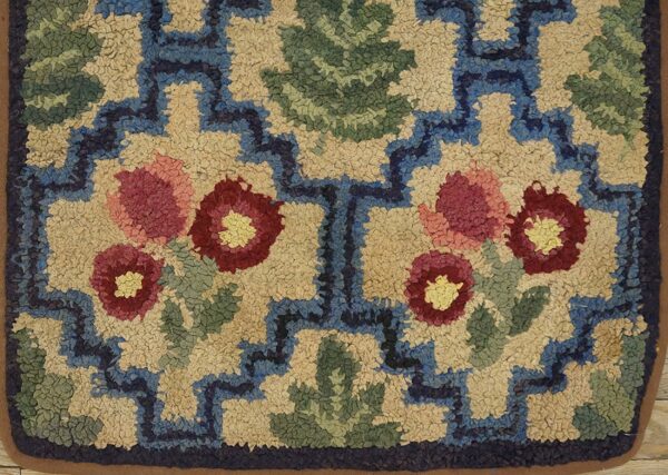 Close-up of a hooked rug featuring stylized red and pink flowers with green leaves against a tan background, bordered by blue geometric shapes. This is the floral hooked rug by rugs on net. By rugs on net