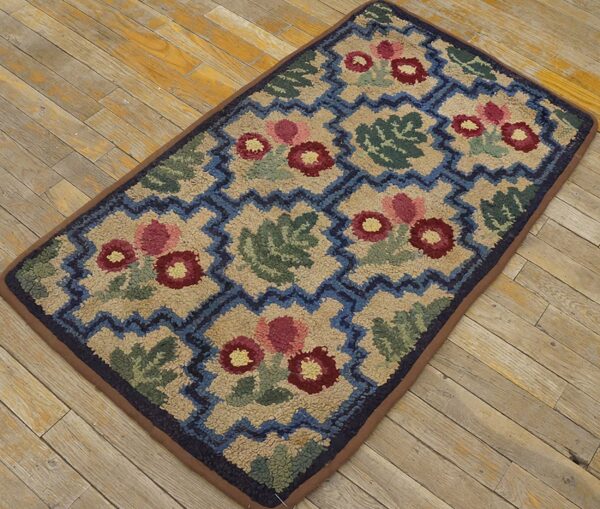 A hooked rug featuring a repeating pattern of stylized red and pink flowers and green leaves set against a beige background with a blue zigzag border, all by rugs on net. By rugs on net