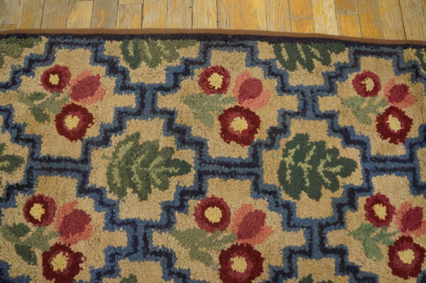 Close-up of a hooked rug featuring stylized red and pink flowers with green leaves against a beige background, framed by a blue geometric border. This is a rugs on net design. By rugs on net