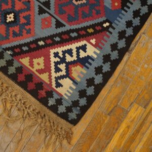 Close-up of a vibrant geometric kilim rug with blue, red, and black patterns, featuring fringed edge on distressed yellow wood floor by Rugs On Net