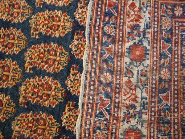 Close-up of a dark blue rug with repeating orange/red motifs next to the reverse side of a patterned rug border by rugs on net
