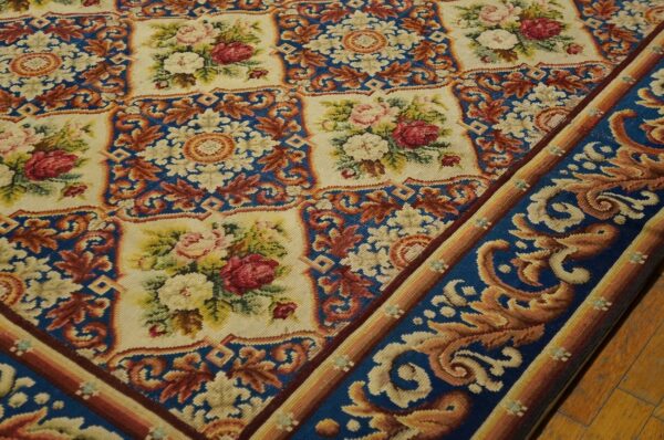 Close-up of a needlepoint rug featuring floral medallions on a cream background framed by a blue border with gold scrolling by rugs on net. By rugs on net