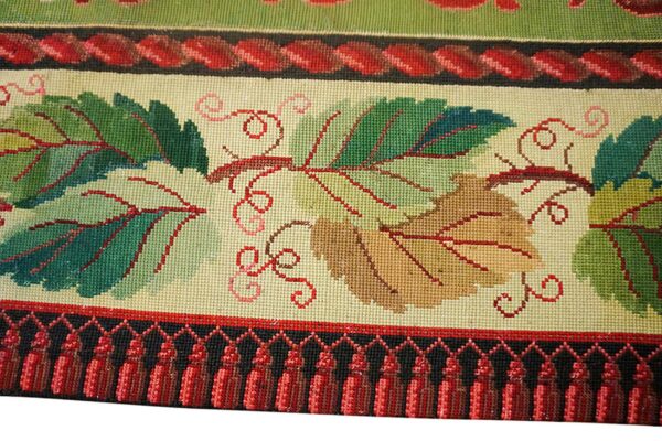 Close-up of a needlepoint rug border featuring green and brown grape leaves with red vines against a cream background, finished with red tassels by rugs on net. By rugs on net