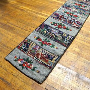 A long, narrow hooked rug with colorful abstract blocks and red floral accents on a gray field, placed on a wooden floor, by Rugs On Net
