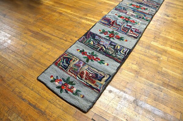 A long, narrow hooked rug with colorful abstract blocks and red floral accents on a gray field, placed on a wooden floor, by rugs on net