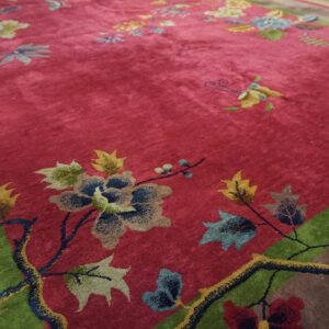 Corner detail of a red Chinese Art Deco rug showing a teal peony, olive and cream leaves and a dark flowering branch against the green inner frame