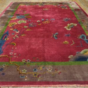 Angled view of a red 1920s Chinese Art Deco carpet on a wood plank floor, showing the taupe border and blue rockwork along the lower edge