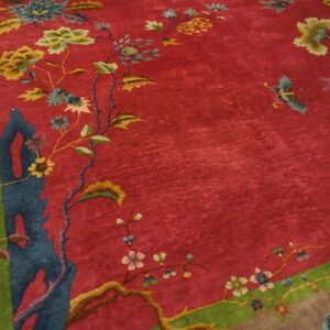 Angled detail of a red Chinese Art Deco rug showing a blue rockwork column and gold and olive florals inside the green inner frame